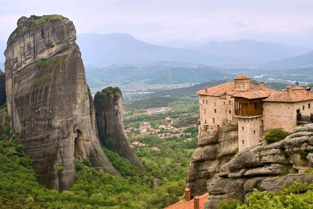 Greece tours: a tall grey rock pillar beside a stone monastery with orange tiled roofs and a wooden balcony on a cliff edge at Meteora, with a green valley and hazy blue mountains behind