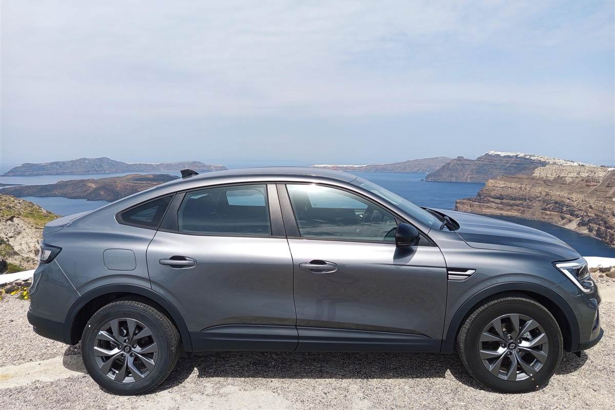 Santorini airport transfer: a grey car parked on a gravel road above the caldera, with the dark volcanic islands, the blue water and the tall brown cliff topped by white houses behind it