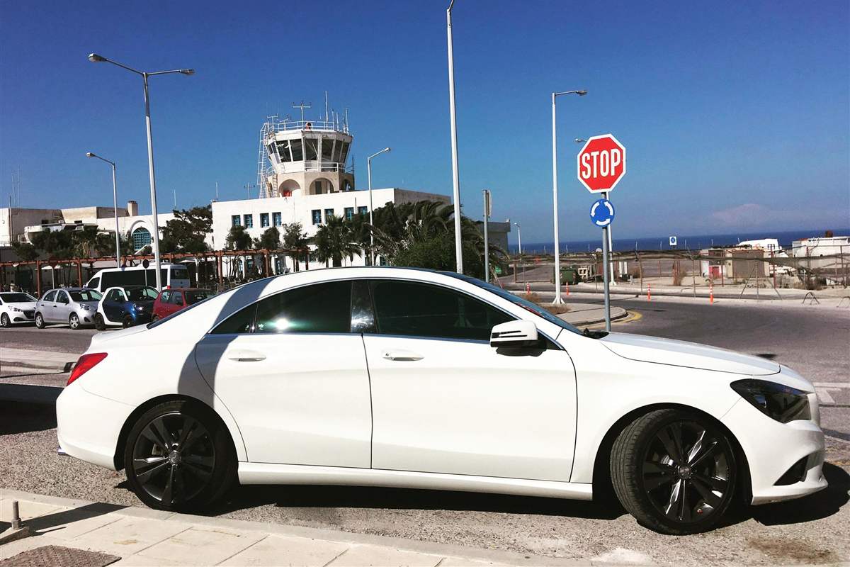Santorini airport transfer: a white saloon car parked on the road outside the Santorini airport terminal, with the white control tower and palm trees behind it under a deep blue sky