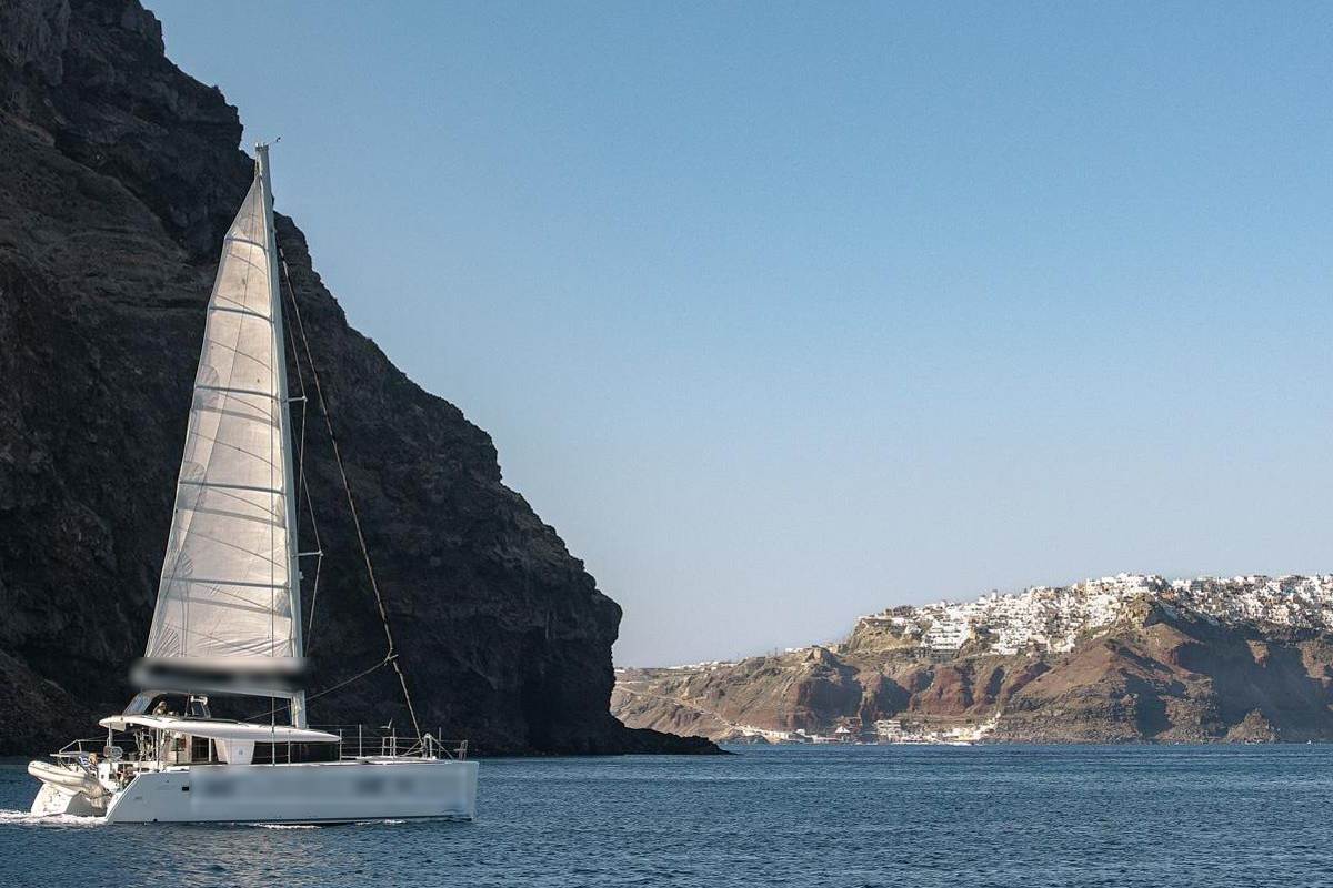 Santorini catamaran cruise: a white catamaran under full sail close to a tall dark volcanic cliff on calm deep blue water, with the white village of Oia on the rim of the caldera in the distance under a clear sky