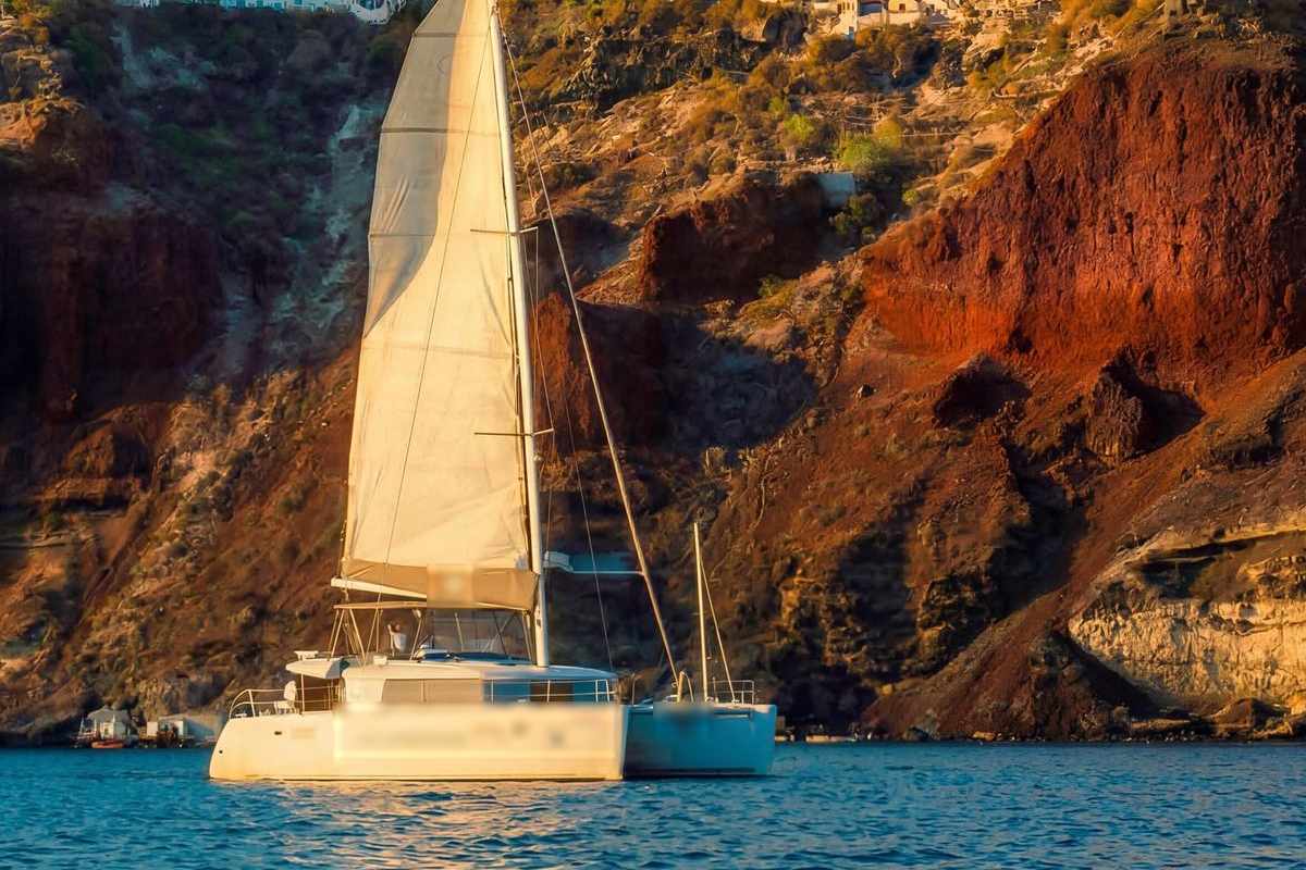 Santorini catamaran cruise: a white catamaran under full sail in golden evening light below a steep red and brown caldera cliff, with a few white houses on the rim far above