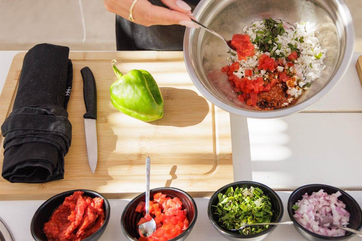 Santorini cooking class: a steel bowl of chopped tomato, rice and herbs being mixed with a spoon, with small bowls of tomato paste, parsley and red onion and a green pepper on a wooden board