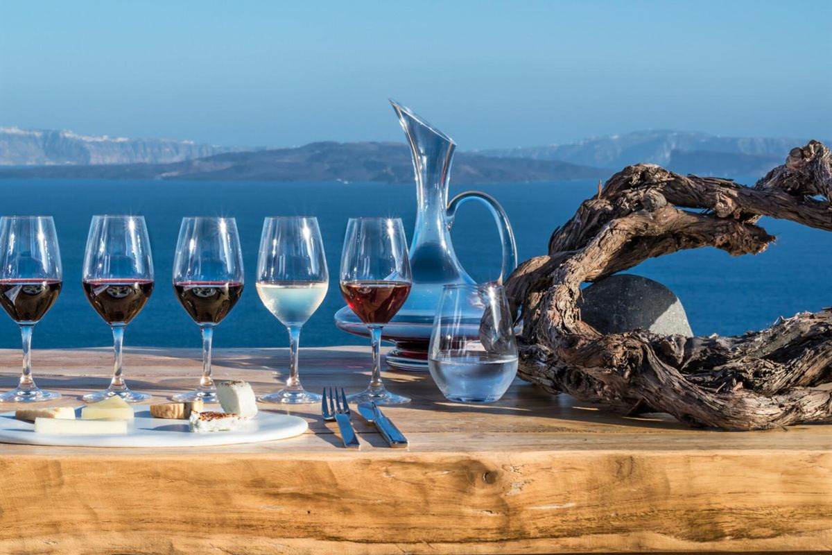 Santorini cooking class: five glasses of red, white and rose wine and a small cheese board on a wooden table beside a twisted old vine, with the deep blue caldera sea and pale cliffs behind under a clear sky