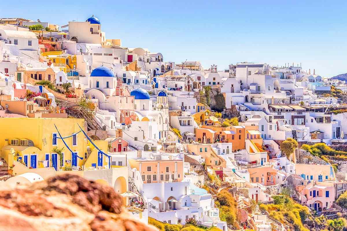 Santorini day trip: the white and pastel houses of Oia stacked on a hillside in bright sun, with three blue church domes among yellow, orange and pink walls under a clear blue sky