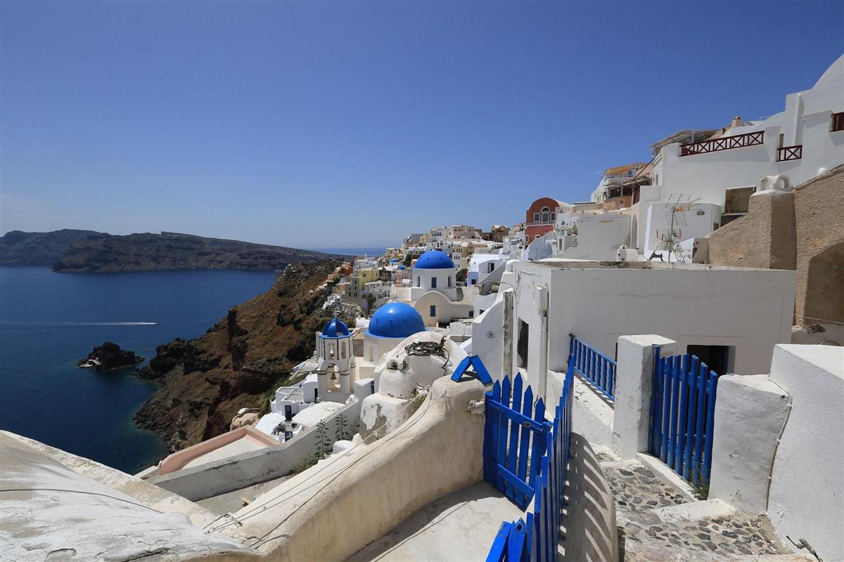 Santorini day trip: blue church domes and a small white bell tower on the caldera rim of Oia above a dark blue bay, with blue wooden gates in the foreground and a red and brown volcanic cliff on the left