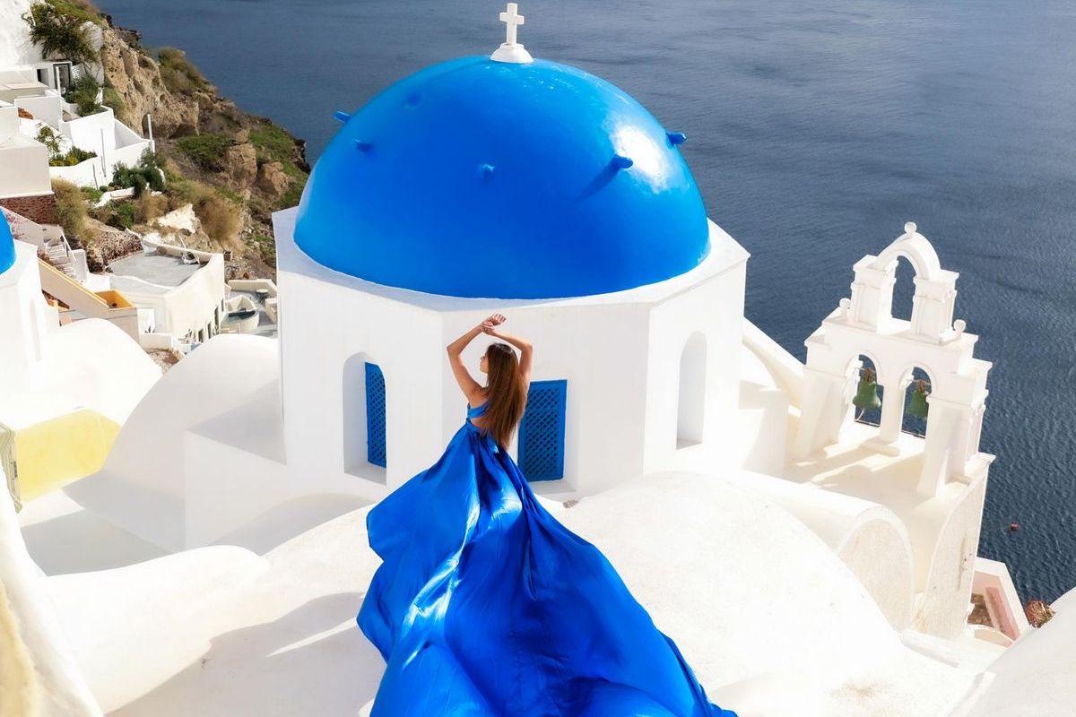 Santorini flying dress photoshoot: a woman seen from behind with her arms raised in a blue dress, its long train flowing over a white roof below the blue dome and bell arches of an Oia church, with the dark blue caldera behind
