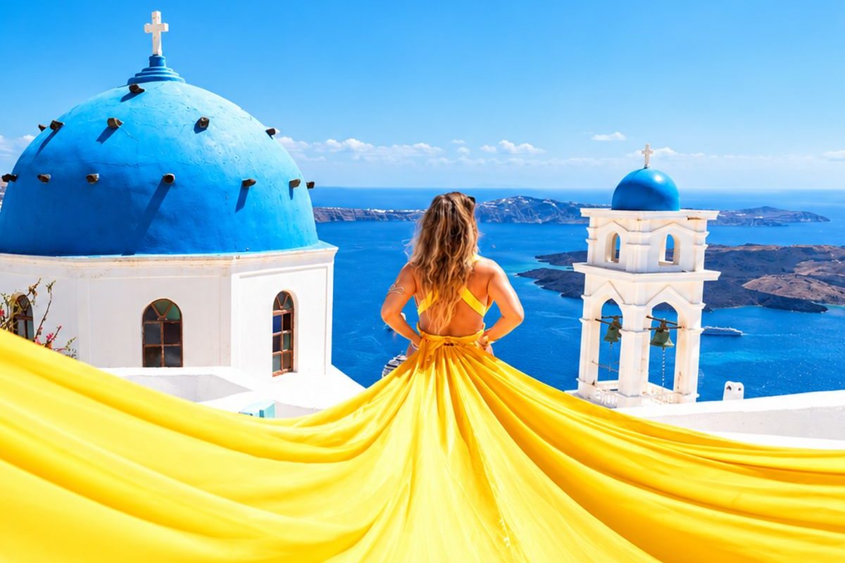 Santorini flying dress photoshoot: a woman seen from behind in a yellow dress, her wide train spread over a white roof in front of a blue dome and a white bell tower, with the blue caldera sea and dark volcanic islands behind