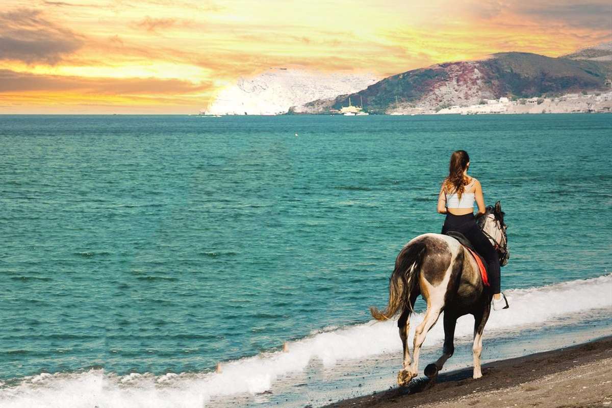 Santorini horseback riding: a rider in a white sun top on a brown, white and black horse walks along the edge of a dark sand beach beside turquoise sea, with a pale pumice headland and a village under an orange sunset sky behind