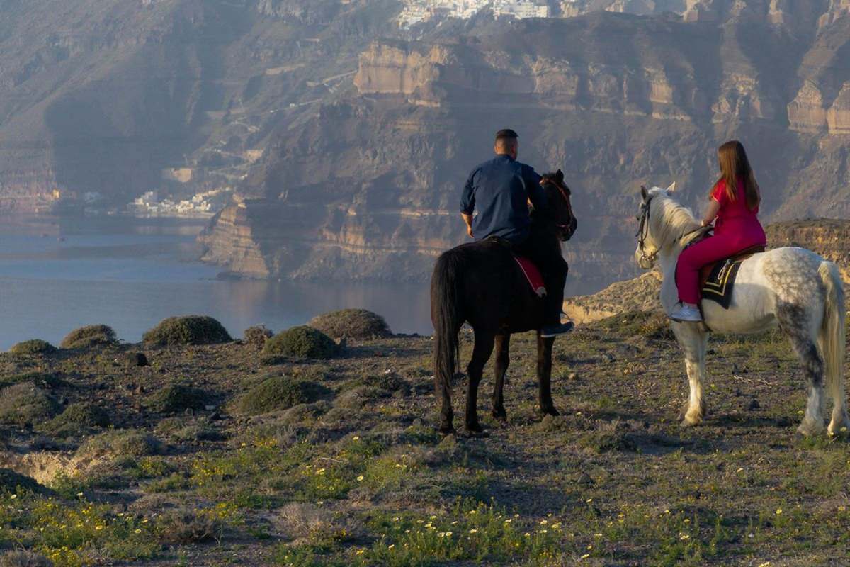 Santorini horseback riding: two riders on a black and a white horse stand on a grassy clifftop with yellow wildflowers, looking across the calm caldera sea to the tall layered cliffs of Santorini with the white houses of Fira on the rim
