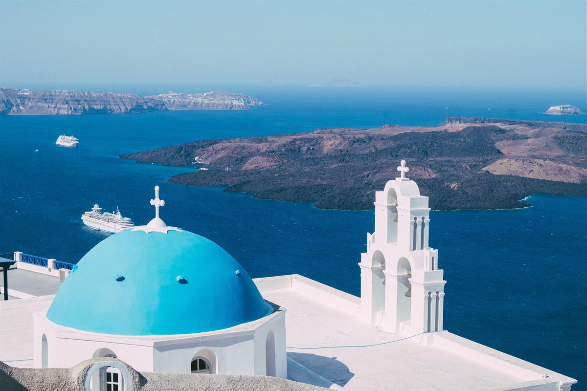 Santorini private tour: a bright blue church dome and a white three-arch bell tower on a whitewashed roof in Fira, with the deep blue caldera, the dark volcanic island of Nea Kameni and two white ships behind