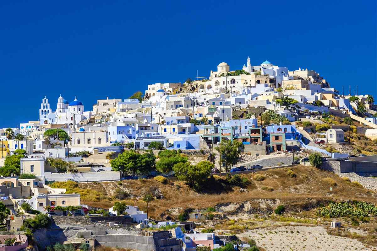 Santorini private tour: a hilltop village of white and pastel houses climbing a dry brown slope, with a white bell tower and blue domes on the left and a deep blue sky above