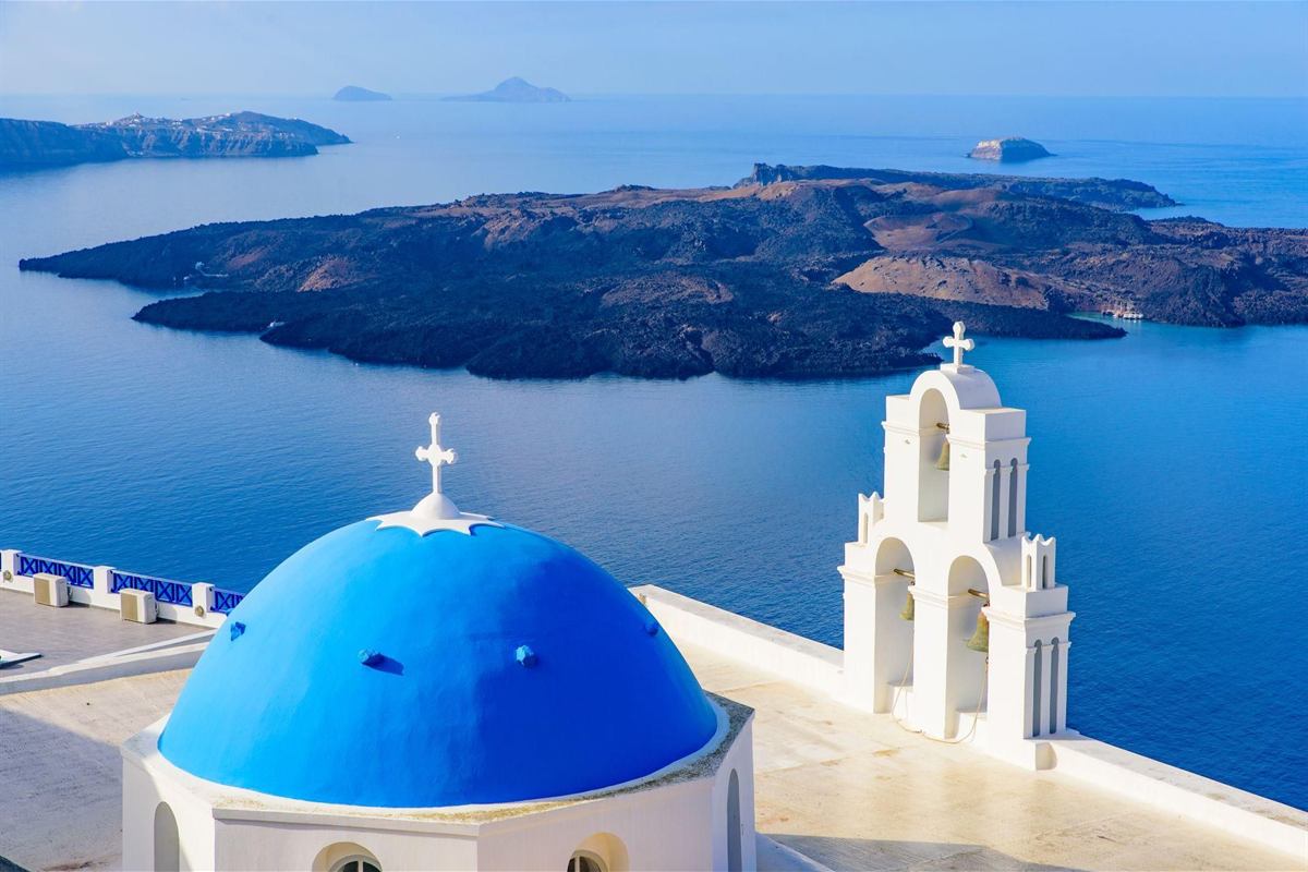 Santorini tours: a blue church dome and a white three-tier bell tower in Fira looking over the calm blue caldera towards the dark volcanic island of Nea Kameni