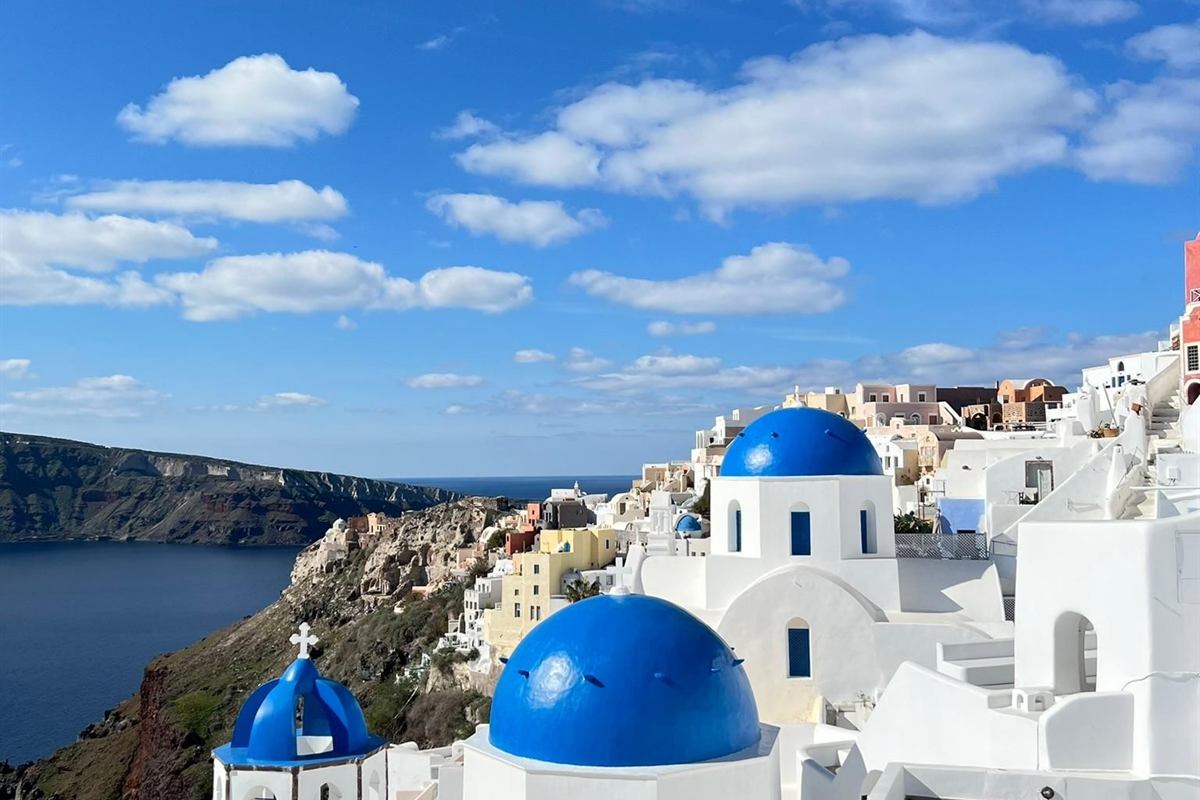 Santorini tours: two bright blue church domes and whitewashed houses of Oia stepping down a rocky cliff towards the deep blue caldera under a sky of white clouds