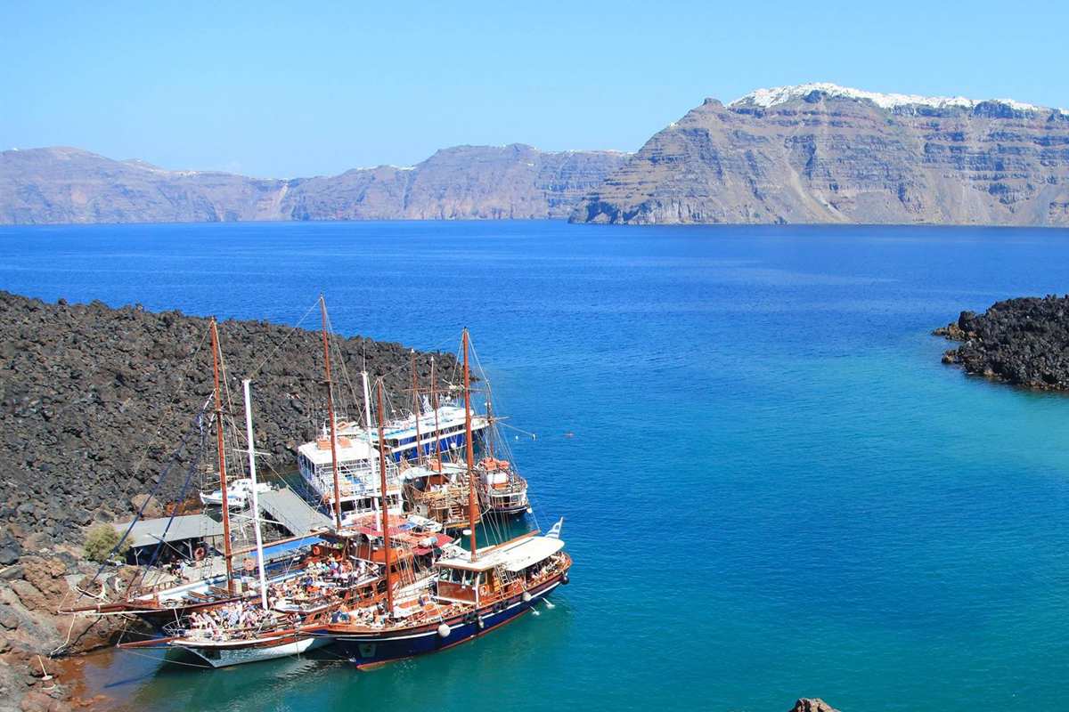 Santorini volcano and hot springs tour: wooden tour boats moored in a black lava cove on Nea Kameni, with the deep blue caldera and the tall Fira cliff behind them in bright sunlight.