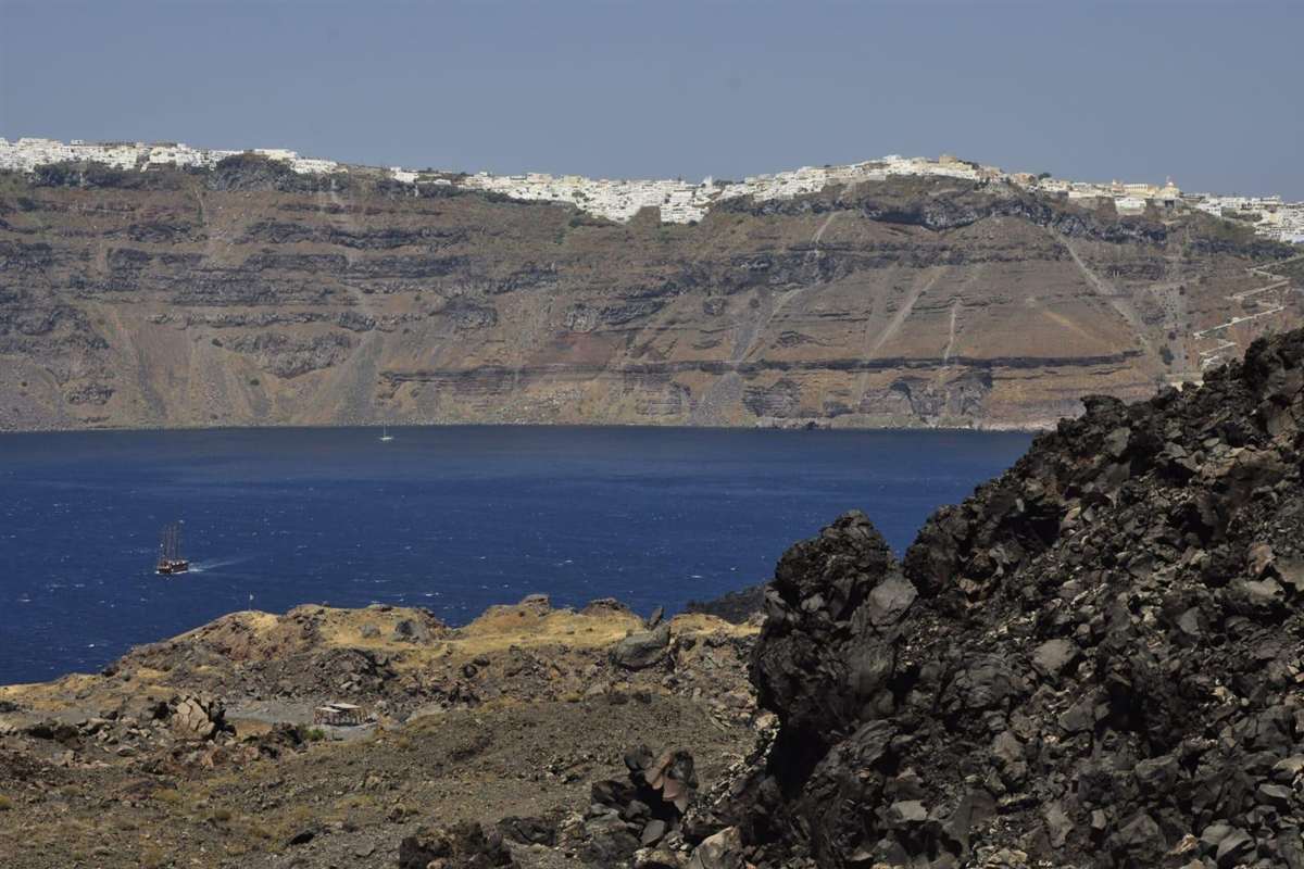 Santorini volcano and hot springs tour: black lava rocks on Nea Kameni in the foreground, a dark blue stretch of the caldera with a small wooden tour boat, and the tall layered cliffs of Santorini topped by the white houses of Fira